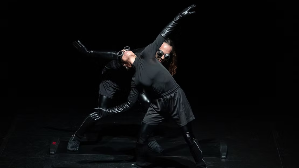 Due performer vestiti completamente di nero, con guanti lucidi e occhiali protettivi, sono in scena su uno sfondo nero. I loro corpi sono intrecciati in una posa dinamica e inclinata, con le braccia tese in direzioni opposte, creando una composizione simmetrica e fluida. L’illuminazione teatrale mette in risalto le forme e i movimenti, mentre il resto dello spazio rimane immerso nel buio.