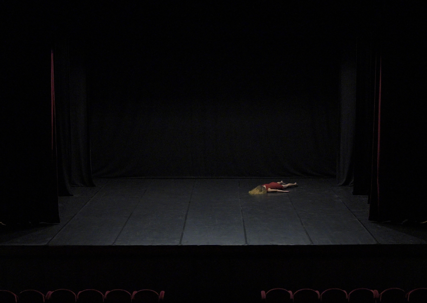 Una danzatrice distesa sul pavimento nero di un palcoscenico teatrale vuoto, illuminata da una luce soffusa. Indossa un vestito rosso e si trova sul lato destro della scena, circondata da tende scure.