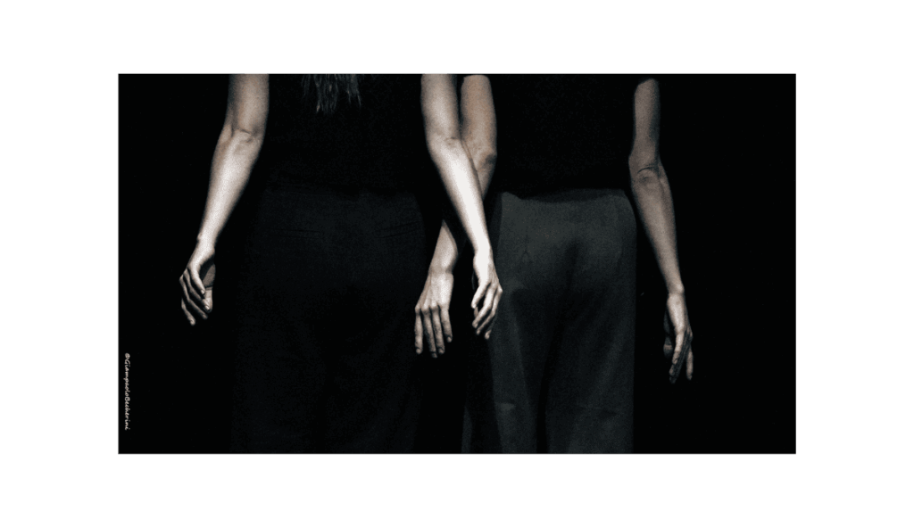 Due figure femminili viste di spalle, illuminate su sfondo nero. Indossano pantaloni scuri e magliette nere, con le braccia rilassate lungo i fianchi. L’inquadratura si concentra sui loro busti e mani, creando un effetto visivo di simmetria e unità