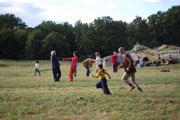 Un gruppo di persone di diverse età partecipa a un'attività all'aperto in un ampio prato circondato da alberi. Alcuni camminano o corrono, altri si tengono per mano, tutti sembrano coinvolti in un momento di gioco o danza collettiva. Sullo sfondo, alcune persone sono sedute vicino a delle rovine in pietra. L'atmosfera è informale e immersa nella natura.