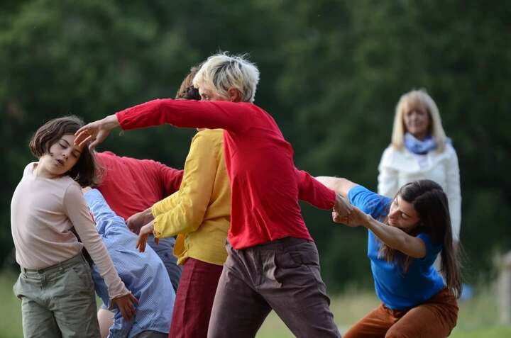 Un gruppo di persone, vestite con abiti colorati, è coinvolto in un'espressione corporea collettiva all’aperto. I loro corpi sono intrecciati in un gesto dinamico e coreografico, con braccia tese e torsioni che suggeriscono un momento di danza o improvvisazione teatrale. Sullo sfondo, una donna osserva la scena con un’espressione serena. La natura verde fa da cornice all’azione.