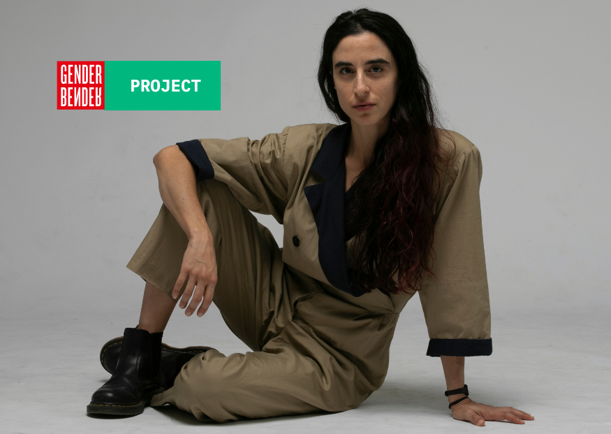Luna Cenere è seduta a terra in posa rilassata, ha lunghi capelli scuri, indossa una tuta beige con dettagli blu scuro e anfibi neri. Sullo sfondo grigio chiaro, in alto a sinistra compare il logo di Gender Bender e la scritta 'PROJECT' su sfondo verde.