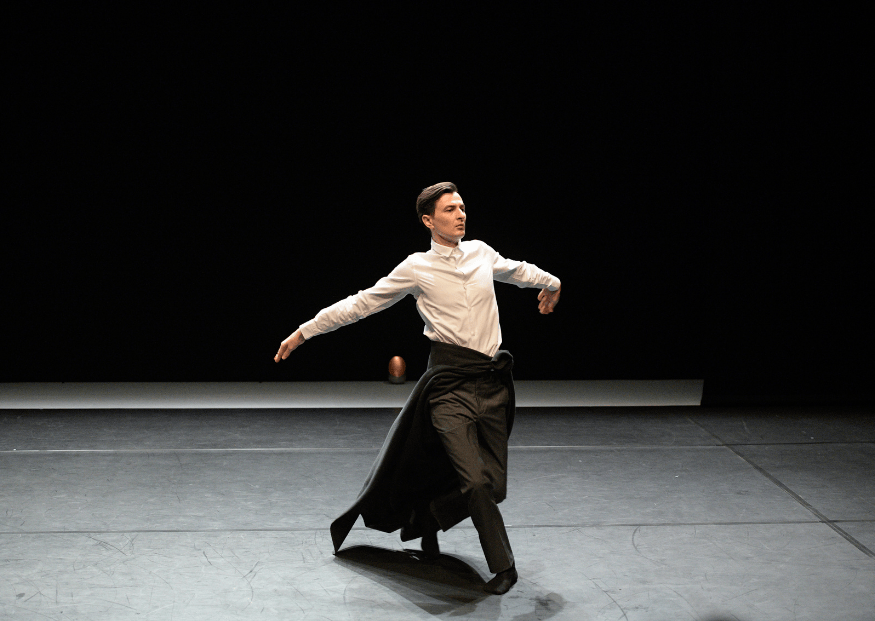 Un danzatore in camicia bianca e pantaloni scuri si muove con eleganza sul palcoscenico, le braccia aperte e il corpo proteso in una rotazione, con il movimento del soprabito che accompagna la danza.