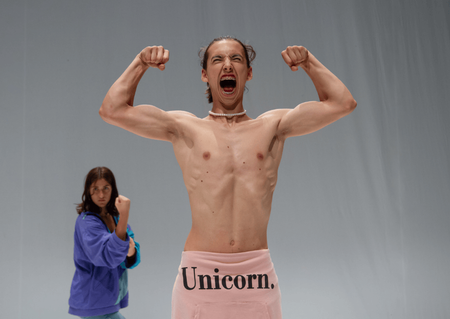 Un performer a torso nudo al centro della scena urla e mostra i muscoli con le braccia sollevate, indossando una felpa rosa legata in vita con la scritta 'Unicorn.'. Sullo sfondo, una persona in abiti casual lo osserva in posa simile.