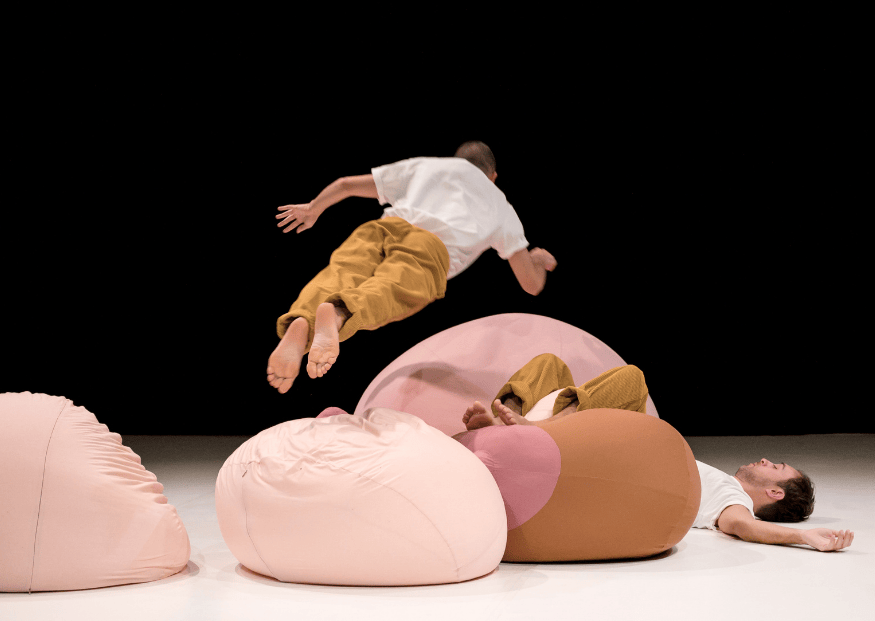 Due performer in scena interagiscono con grandi cuscini colorati: uno salta in aria a braccia aperte mentre l’altro è disteso a terra, creando un’atmosfera giocosa e dinamica.