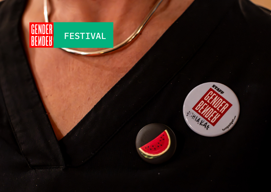 Primo piano di una persona con una collana d’argento e due spille appuntate sulla maglia nera, una con il logo rosso e bianco di Gender Bender Festival e l’altra con una fetta d’anguria stilizzata.