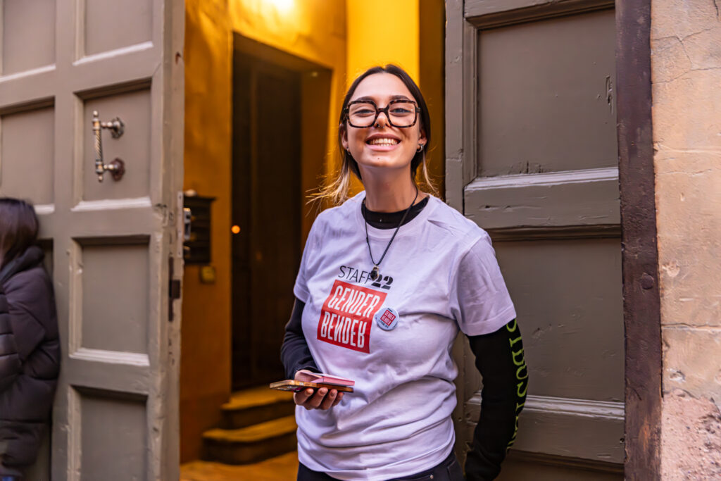 Una giovane volontaria sorridente, con occhiali e capelli raccolti, è appoggiata all’ingresso di un edificio con una porta aperta alle sue spalle illuminata da una luce calda. Indossa una maglietta bianca con la scritta “Staff Gender Bender” e tiene uno smartphone in mano, accogliendo i visitatori.